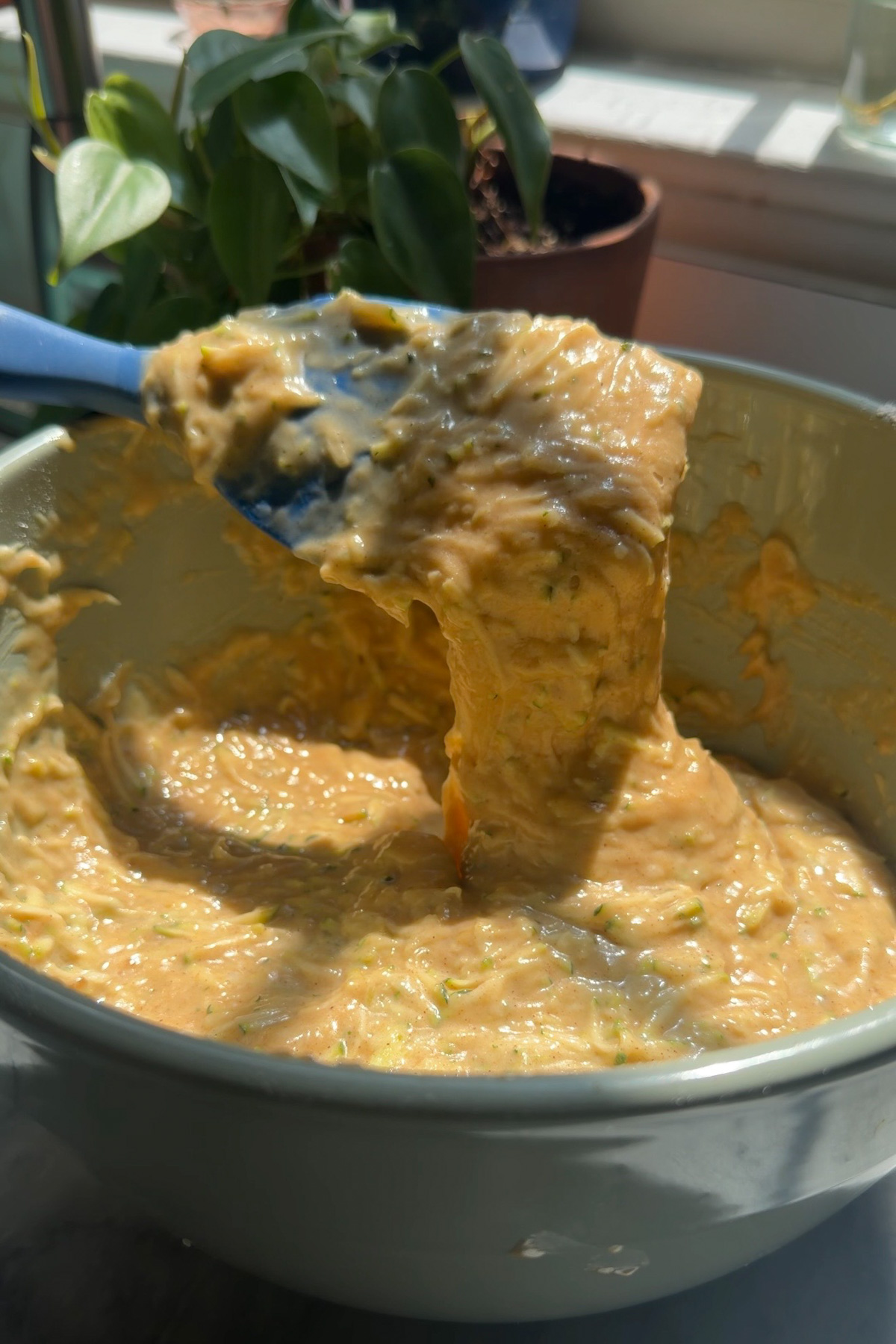 The batter for sourdough discard zucchini muffins in a ceramic mixing bowl with a plant in the background.