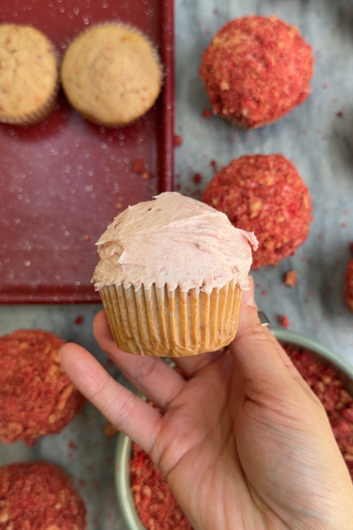 Hand holding a strawberry crunch cupcake with more cupcakes in the background.