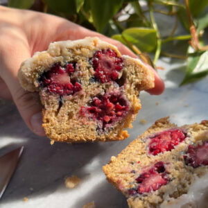 A hand holding a half of a banana blackberry muffin cut in half, showing the berries inside with the other half lying on the counter next to it.