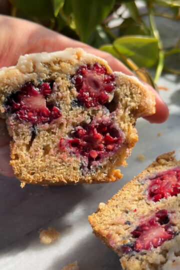 A hand holding a half of a banana blackberry muffin cut in half, showing the berries inside with the other half lying on the counter next to it.