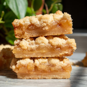 Three apple pie shortbread crumb bars stacked on top of each other in front of a green plant on a marble countertop.