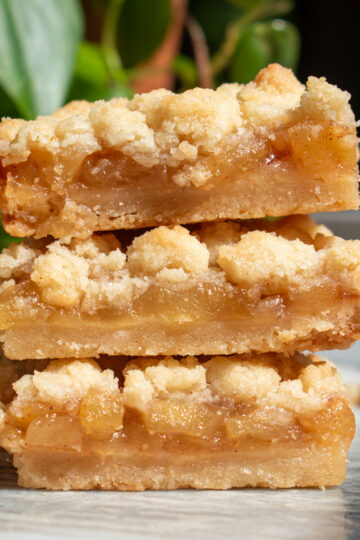 Three apple pie shortbread crumb bars stacked on top of each other in front of a green plant on a marble countertop.