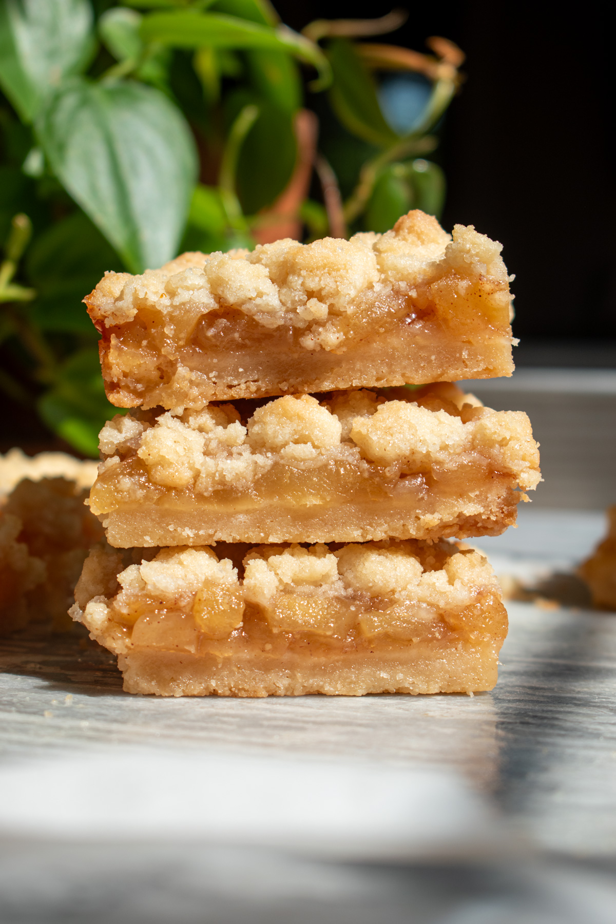 Three apple pie shortbread crumb bars stacked on top of each other in front of a green plant on a marble countertop.