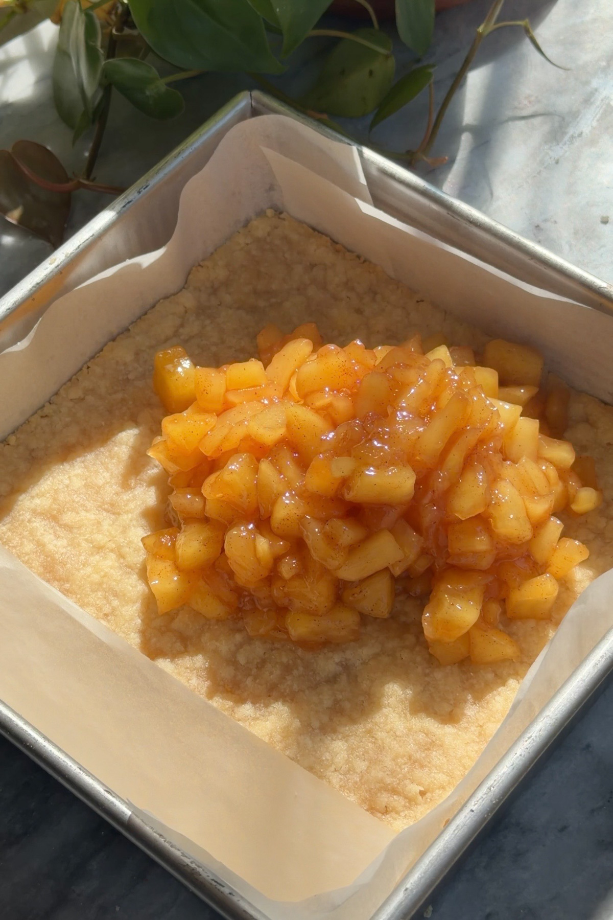 Apple pie filling being added to a pre baked crust in a lined 8x8 baking pan for apple pie crumb bars.