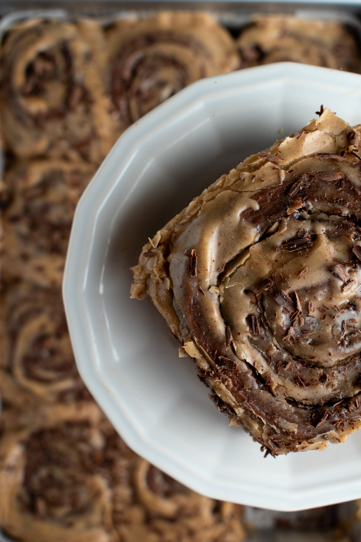 Overhead picture of a chocolate cinnamon roll with cinnamon espresso cream cheese icing on a small plate with other cinnamon rolls in the background. 