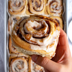Overhead picture of a hand holding an apple pie cinnamon roll with more cinnamon rolls in the background.