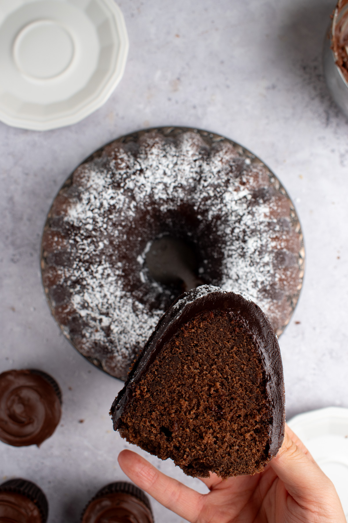 Picture of a hand holding a slice of chocolate sour cream pound cake with a bundt cake and cupcakes in the background.