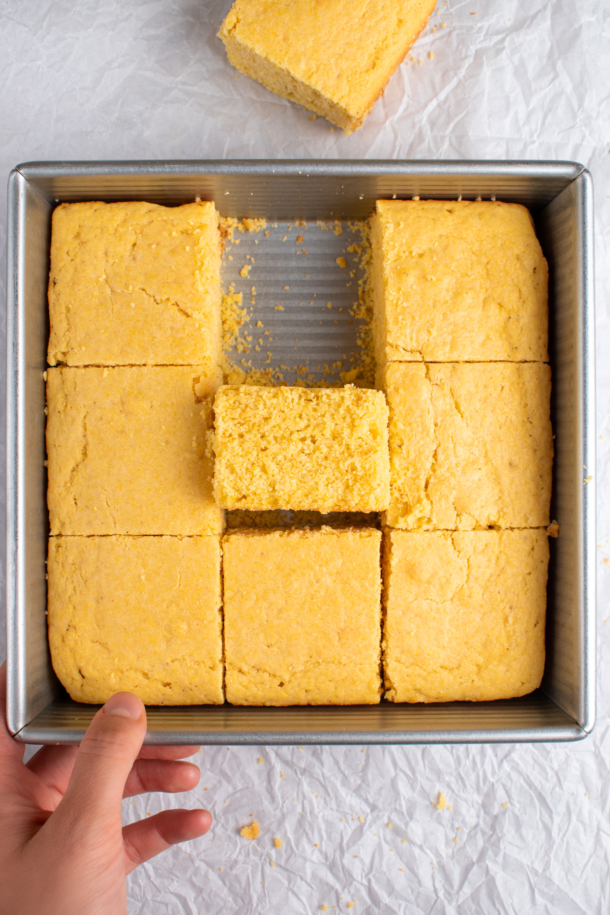 Picture of sourdough cornbread cut into 9 pieces in an 8x8 pan. One piece is out side of the pan and there is a hand holding the bottom of the pan.