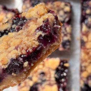 Picture of a blueberry crumb bar with a bite taken out and more crumb bars in the background on parchment paper.