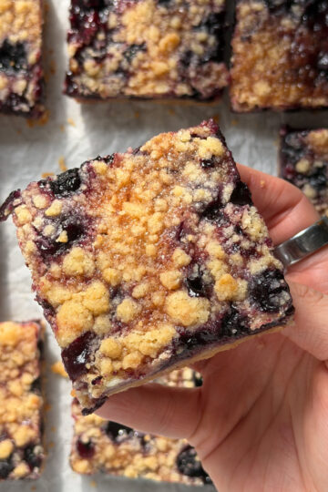 Overhead picture of a right hand holding a blueberry crumb bar above other crumb bars on parchment paper.