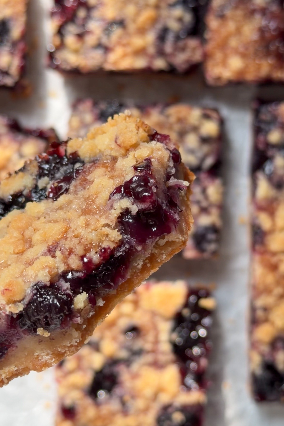 Overhead view of a blueberry crumb bar with a bite taken out, held up in the air against a background of other crumb bars on white parchment paper.
