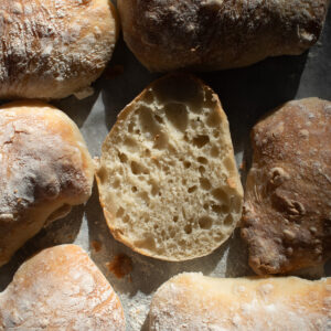 picture of a sourdough discard ciabatta roll cut in half surrounded by other rolls on a lined baking sheet.