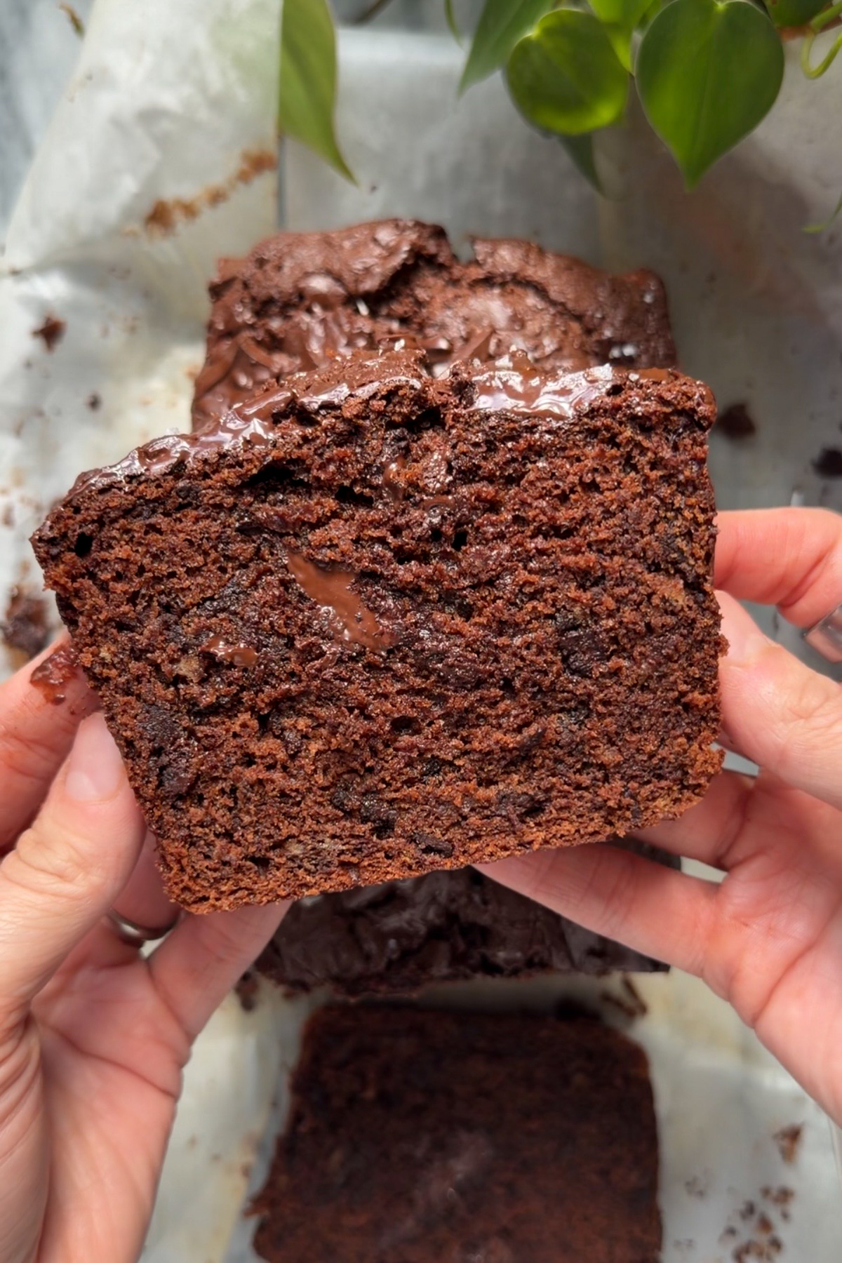 Overhead picture of two hands holding a slice of chocolate chunk banana bread with the rest of the loaf below on parchment paper.