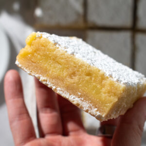Overhead picture of a hand holding a lemon crumb bar showing its layers with more lemon crumb bars laying flat in the background.