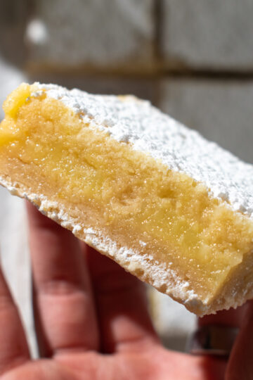 Overhead picture of a hand holding a lemon crumb bar showing its layers with more lemon crumb bars laying flat in the background.