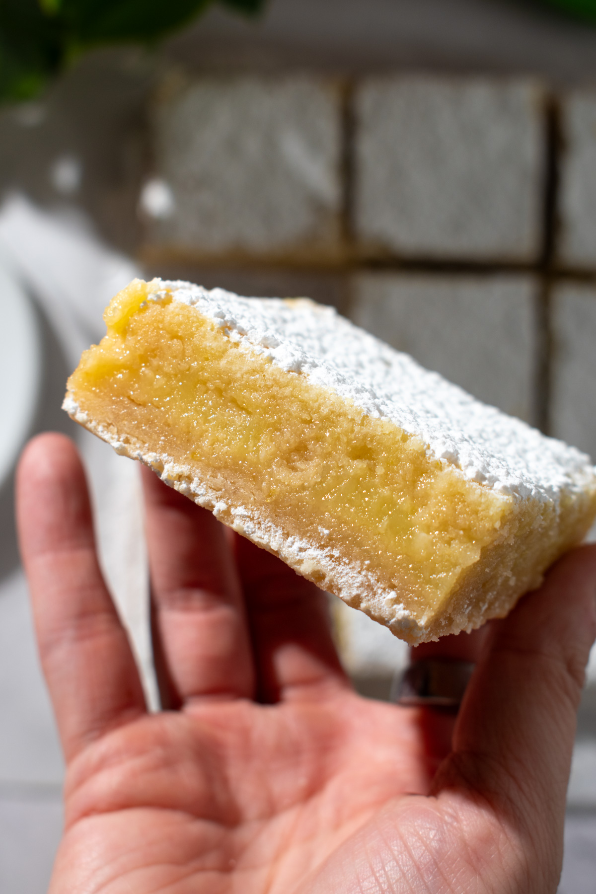 Overhead picture of a hand holding a lemon crumb bar showing its layers with more lemon crumb bars lying flat in the background.