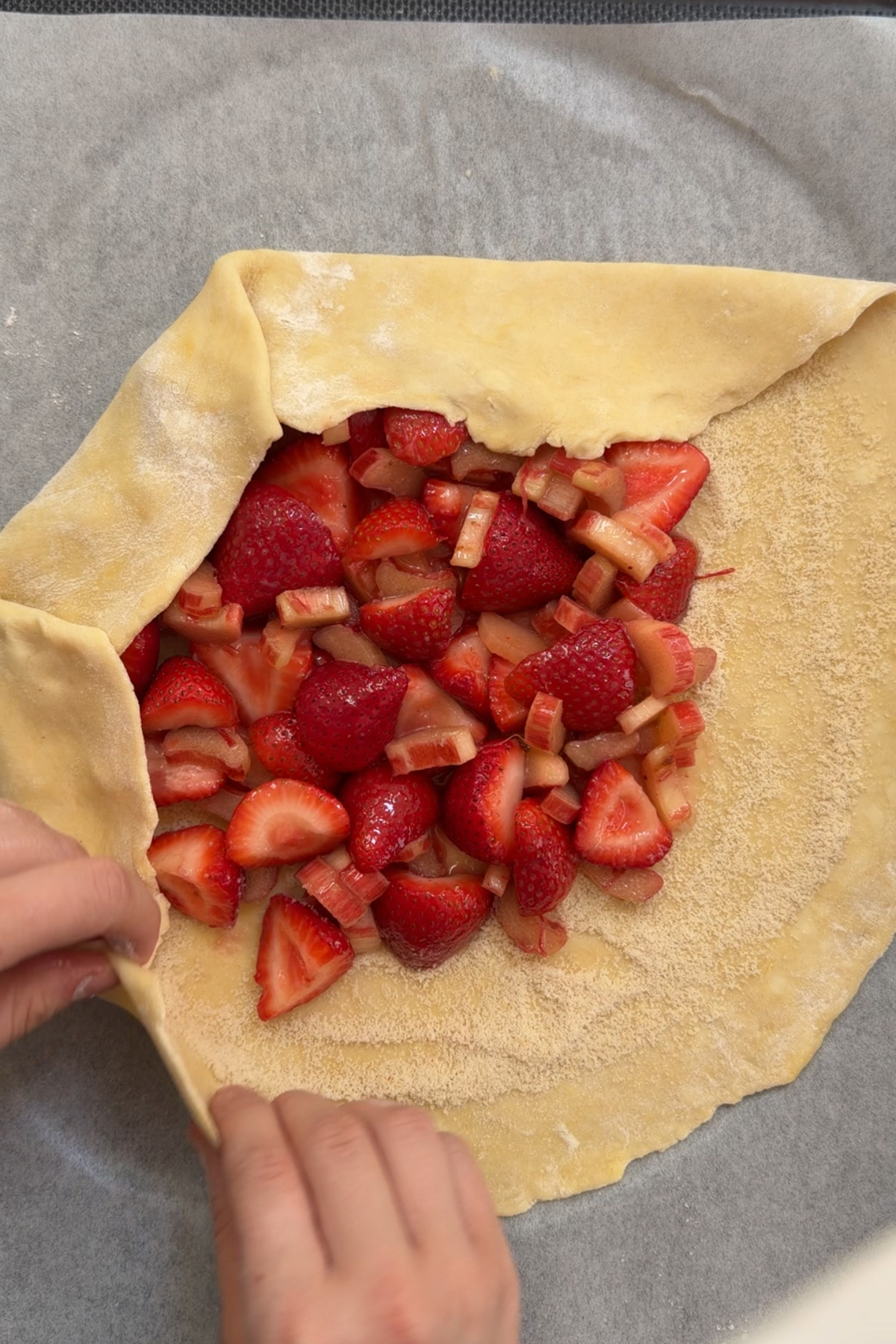 Overhead picture of two hands shaping and folding a strawberry-rhubarb galette over the filling before baking.