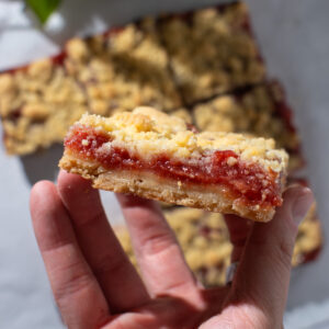 A hand holding strawberry rhubarb crumb bars, showing the layers of crust and jam with more crumb bars laying flat in the background.