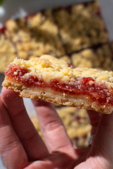 A hand holding strawberry rhubarb crumb bars, showing the layers of crust and jam with more crumb bars laying flat in the background.