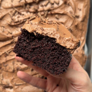 A hand holding a slice of chocolate cream cheese sheet cake to show the texture with the remaining cake lying flat in the background.