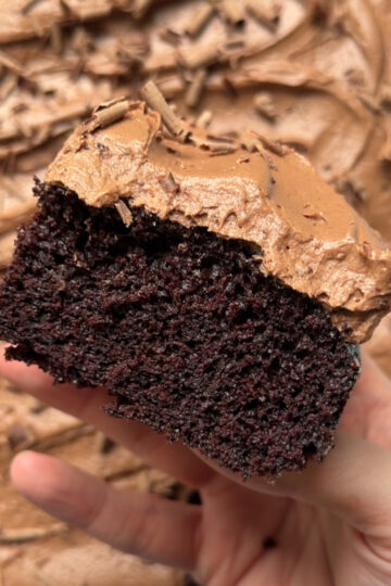 A hand holding a slice of chocolate cream cheese sheet cake to show the texture with the remaining cake lying flat in the background.