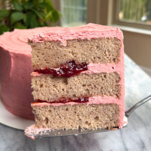 A slice of a three-layer strawberry cake with strawberry filling with the rest of the cake in the background in front of a green plant and windows.
