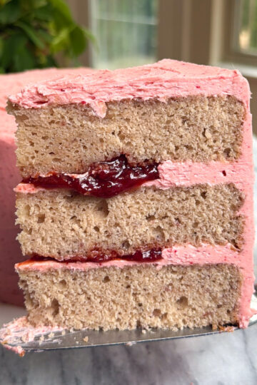 A slice of a three-layer strawberry cake with strawberry filling with the rest of the cake in the background in front of a green plant and windows.