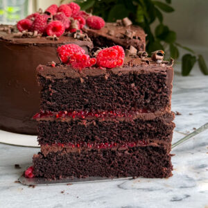 A slice of dark chocolate and raspberry cake with chocolate ganache frosting and raspberry compote between the layers. The cake is topped with chocolate curls and fresh raspberries.