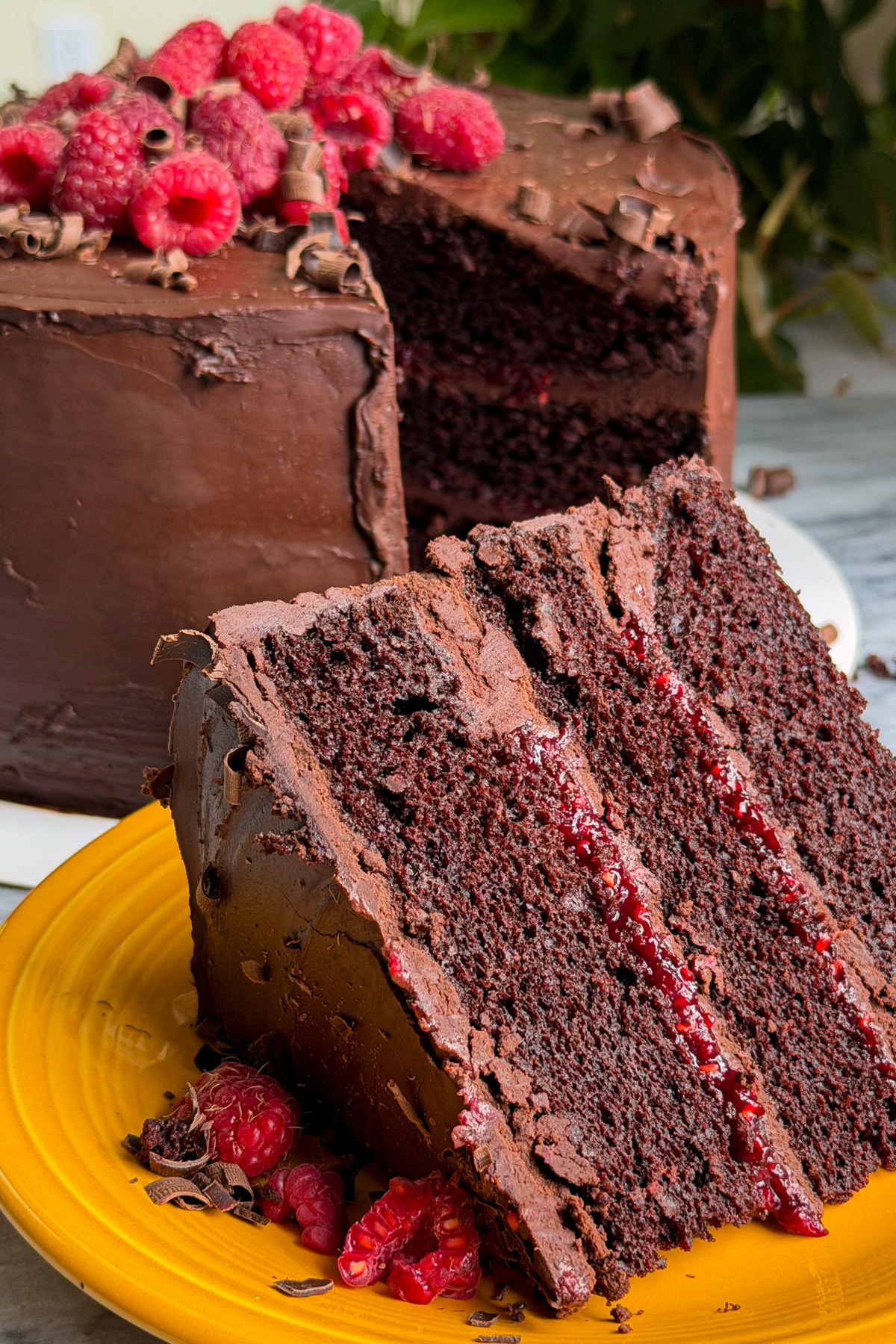 A slice of dark chocolate and raspberry cake with chocolate ganache frosting and raspberry compote between the layers on a yellow plate in front of the rest of the cake. The cake is topped with chocolate curls and fresh raspberries.