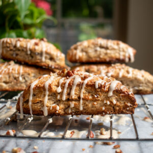 A maple pecan scone on a cooling rack covered in maple glaze and topped with chopped pecans, with more scones stacked on top of each other in the background.