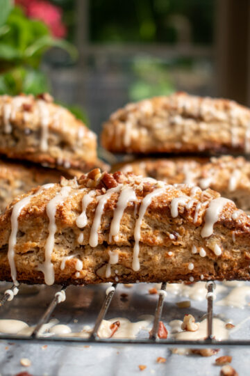 A maple pecan scone on a cooling rack covered in maple glaze and topped with chopped pecans, with more scones stacked on top of each other in the background.