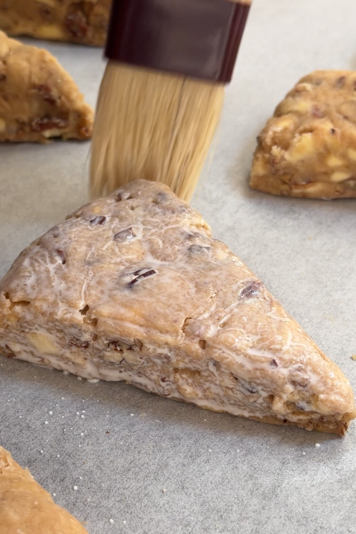 A maple pecan scone on a baking sheet being brushed with heavy cream before baking. 