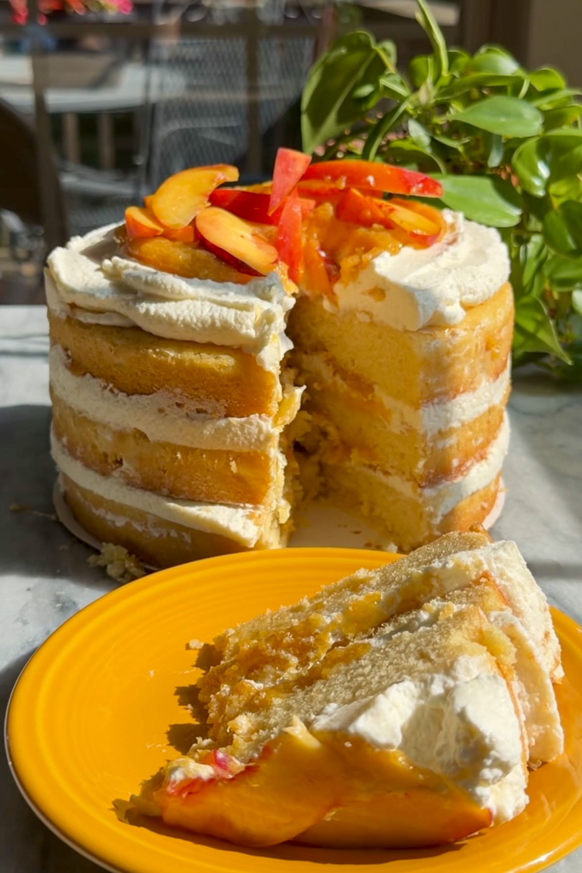 A slice of brown butter bourbon peaches and cream cake with layers of whipped cream and bourbon peach compote on a plate in front of the rest of the cake.
