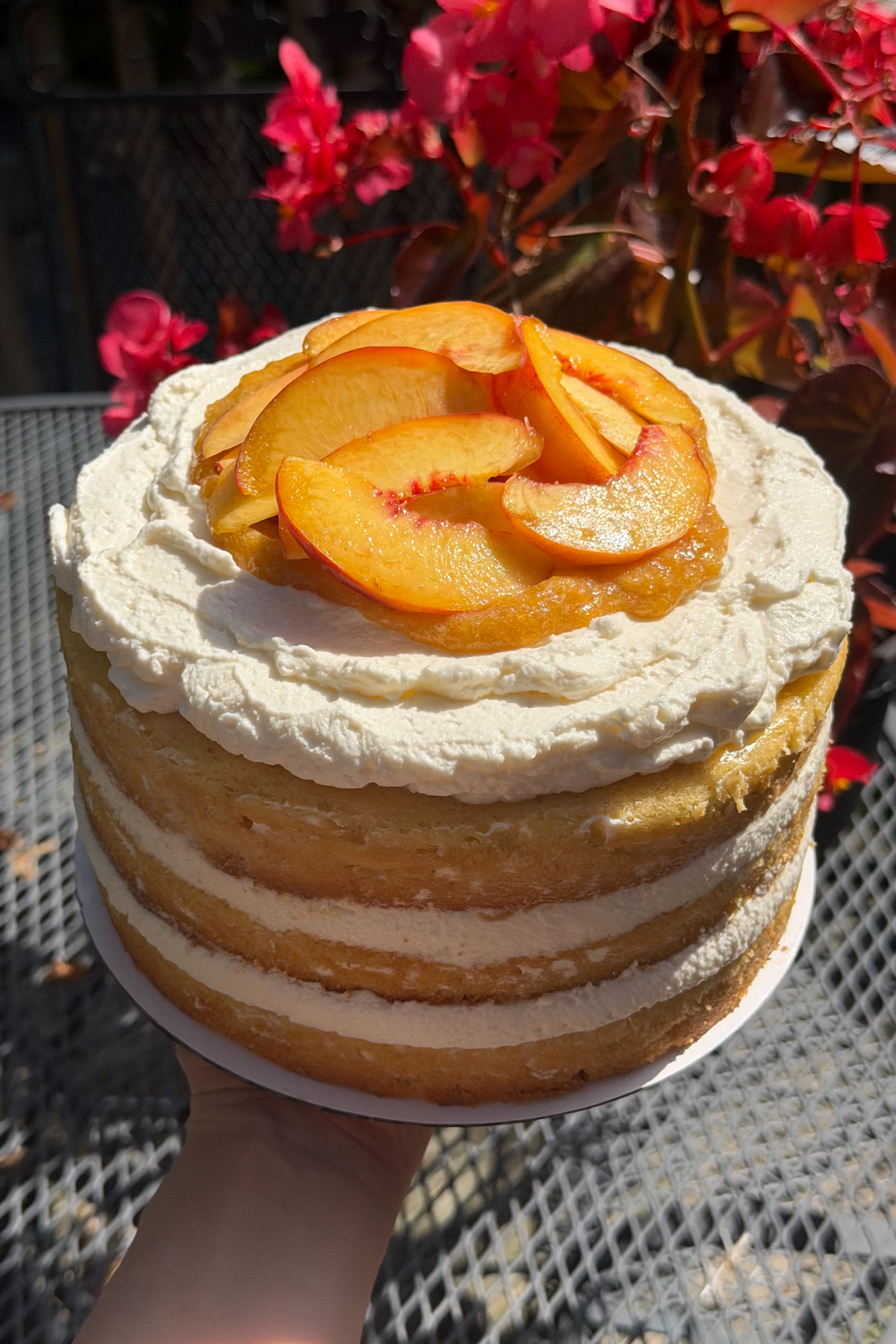 A brown butter bourbon peaches and cream cake being held up in front of pink flowers.