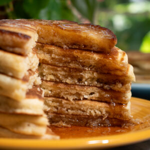 Six sourdough discard pancakes stacked on a yellow plate in front of a window with a slice taken out of it to show the layers and maple syrup drip.