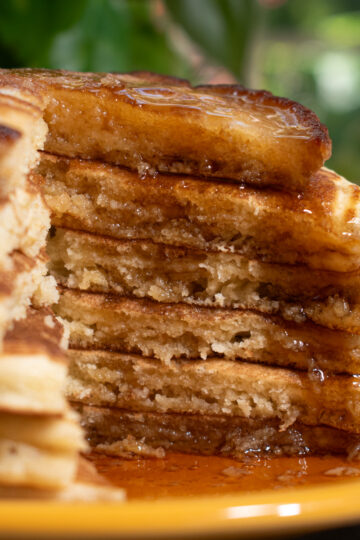 Six sourdough discard pancakes stacked on a yellow plate in front of a window with a slice taken out of it to show the layers and maple syrup drip.