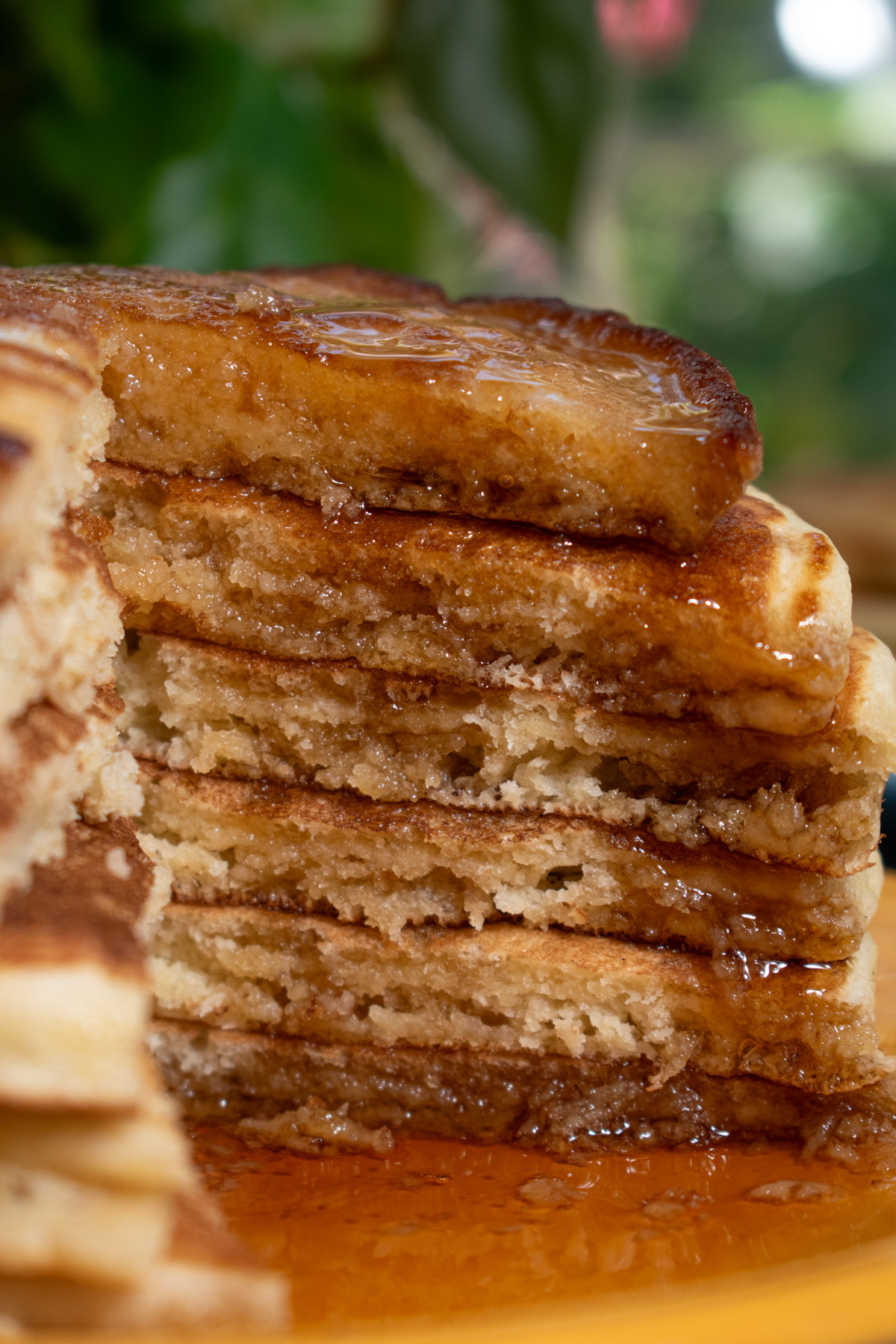 Six sourdough discard pancakes stacked on a yellow plate in front of a window with a slice taken out of it to show the layers and maple syrup drip.