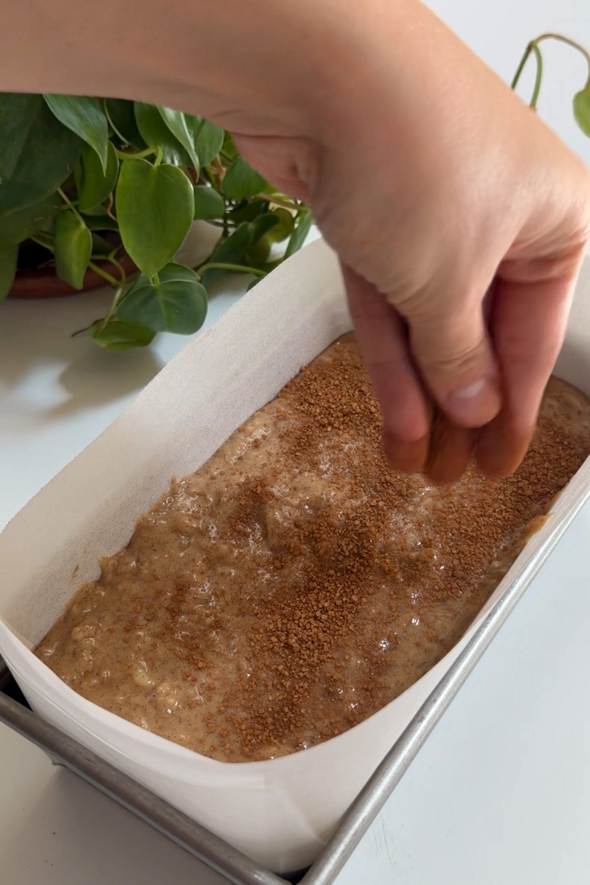 Apple butter banana bread batter in a 9x5 lined loaf pan with a hand sprinkling cinnamon sugar on top.