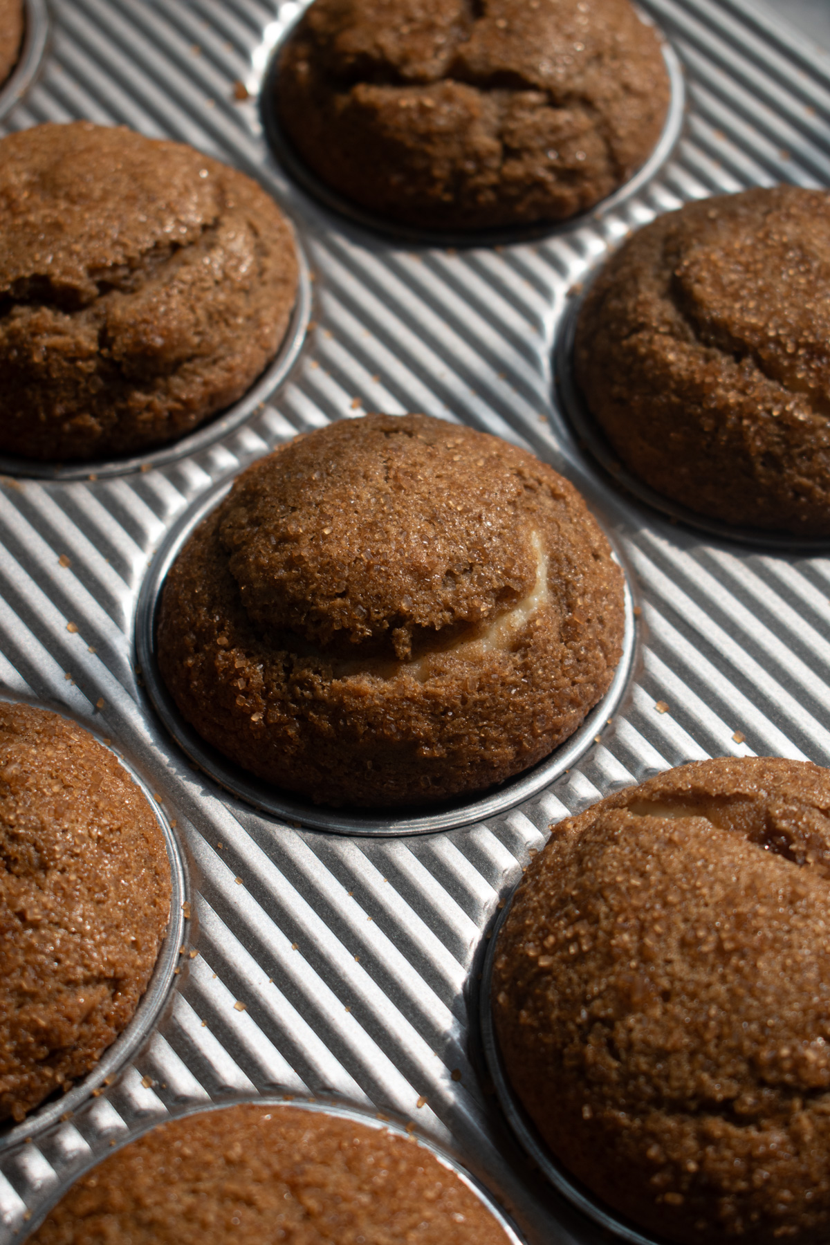 Baking cheesecake-stuffed apple butter muffins in a muffin pan after coming out of the oven with visible cream cheese swirls and crinkly sugar-coated domed tops.