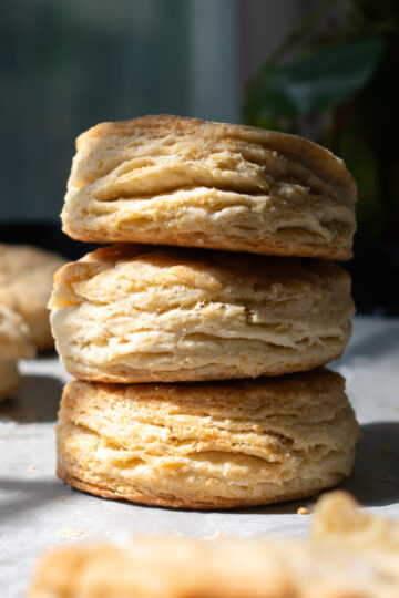 Three golden flaky sourdough discard biscuits stacked on top of each other on a parchment lined baking sheet surrounded by more biscuits.