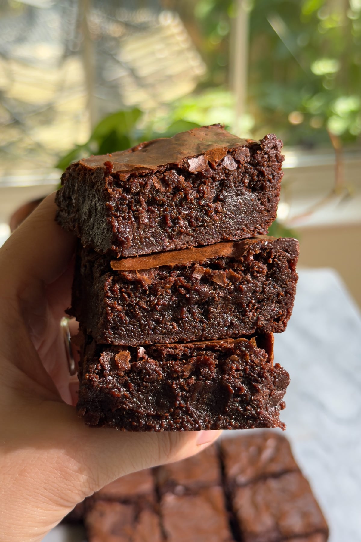 A hand holding three crackly fudgy brownies stacked on top of each other in front of a window.