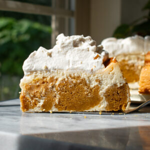 A slice of pumpkin cake cheesecake with brown sugar whipped cream on a marble countertop in front of a window.