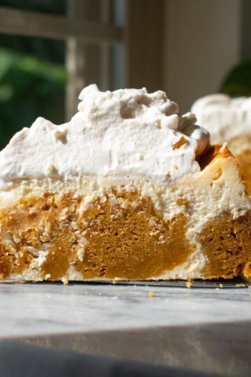A slice of pumpkin cake cheesecake with brown sugar whipped cream on a marble countertop in front of a window.