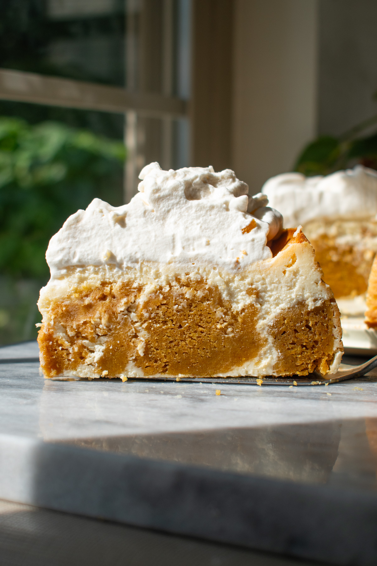 A slice of pumpkin cake cheesecake with brown sugar whipped cream on a marble countertop in front of a window.