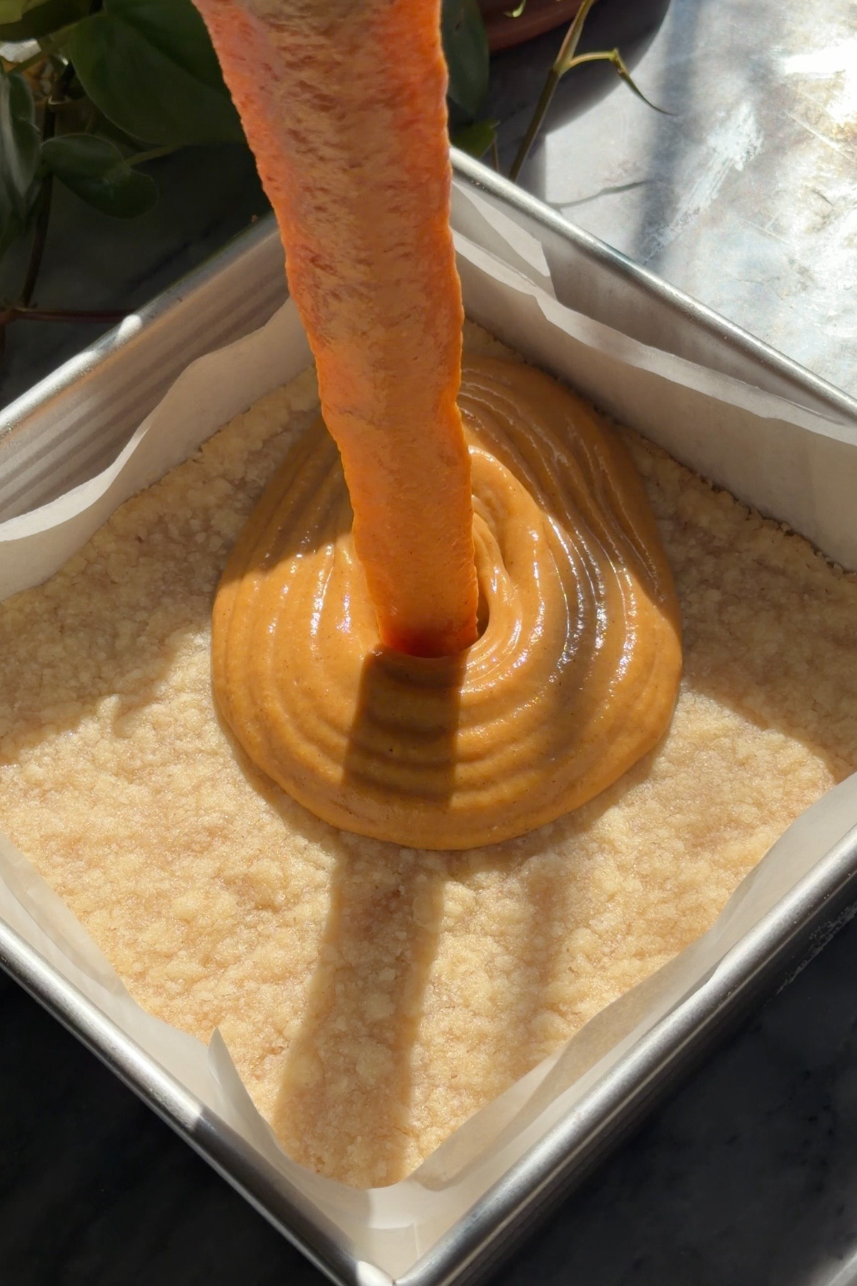Pumpkin pie filling pouring onto a par-baked shortbread crust for pumpkin pie crumb bars in a lined 8x8 baking pan.