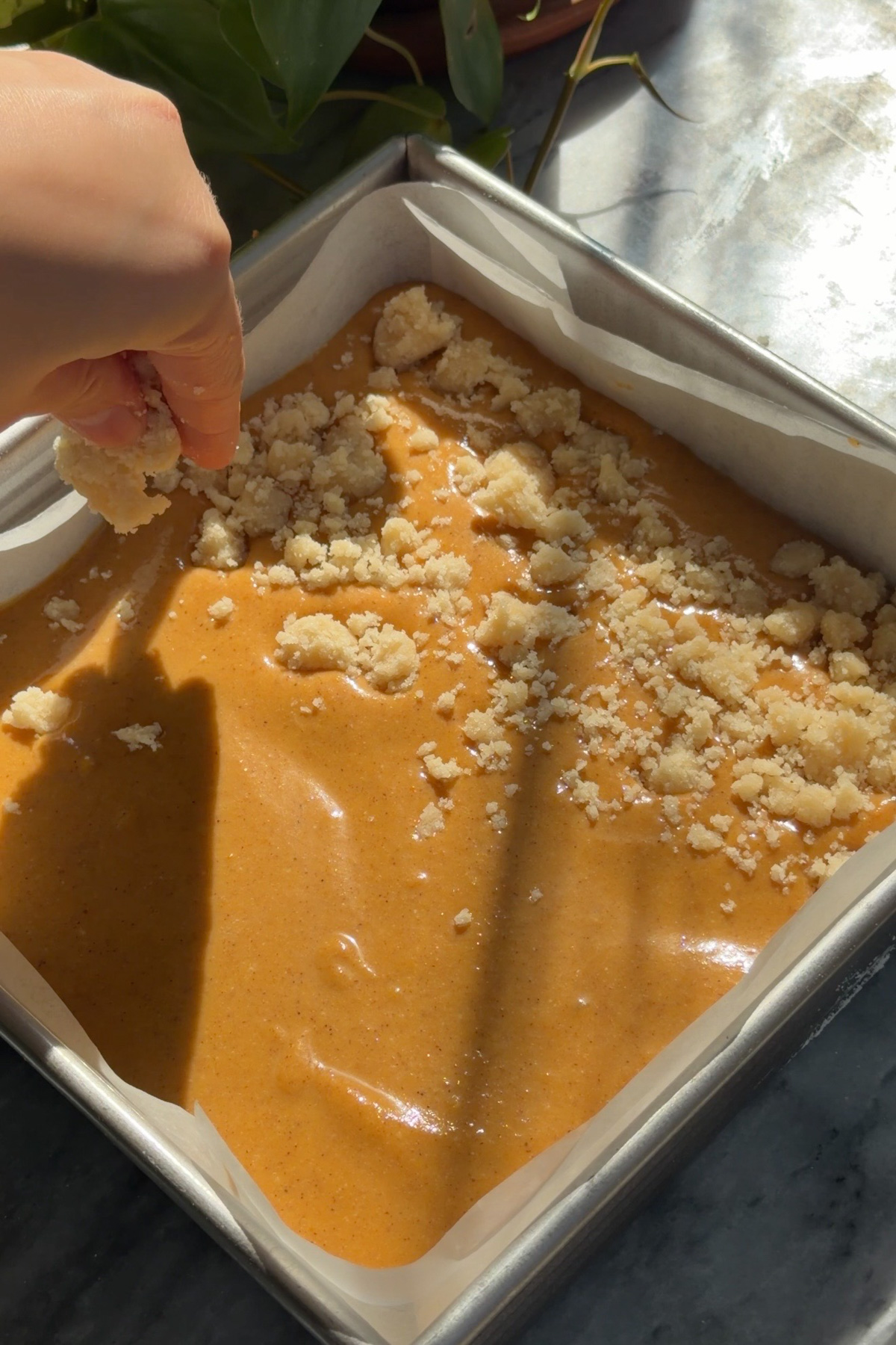 Shortbread crumble sprinkling on top of pumpkin pie filling for pumpkin pie crumb bars in a lined 8x8 baking pan.
