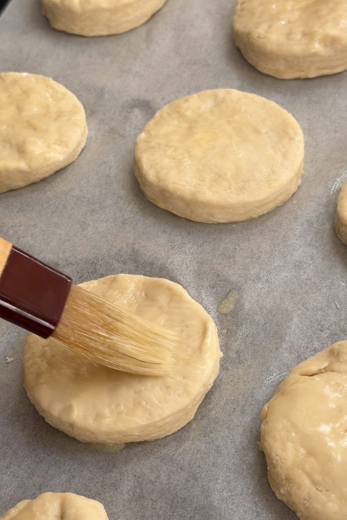 Sourdough discard biscuits are brushed with butter on a lined baking sheet before baking.