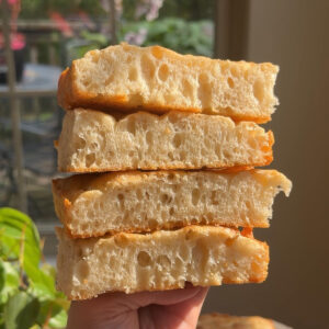 A hand holding four slices of sourdough discard focaccia in front of plants and a window.