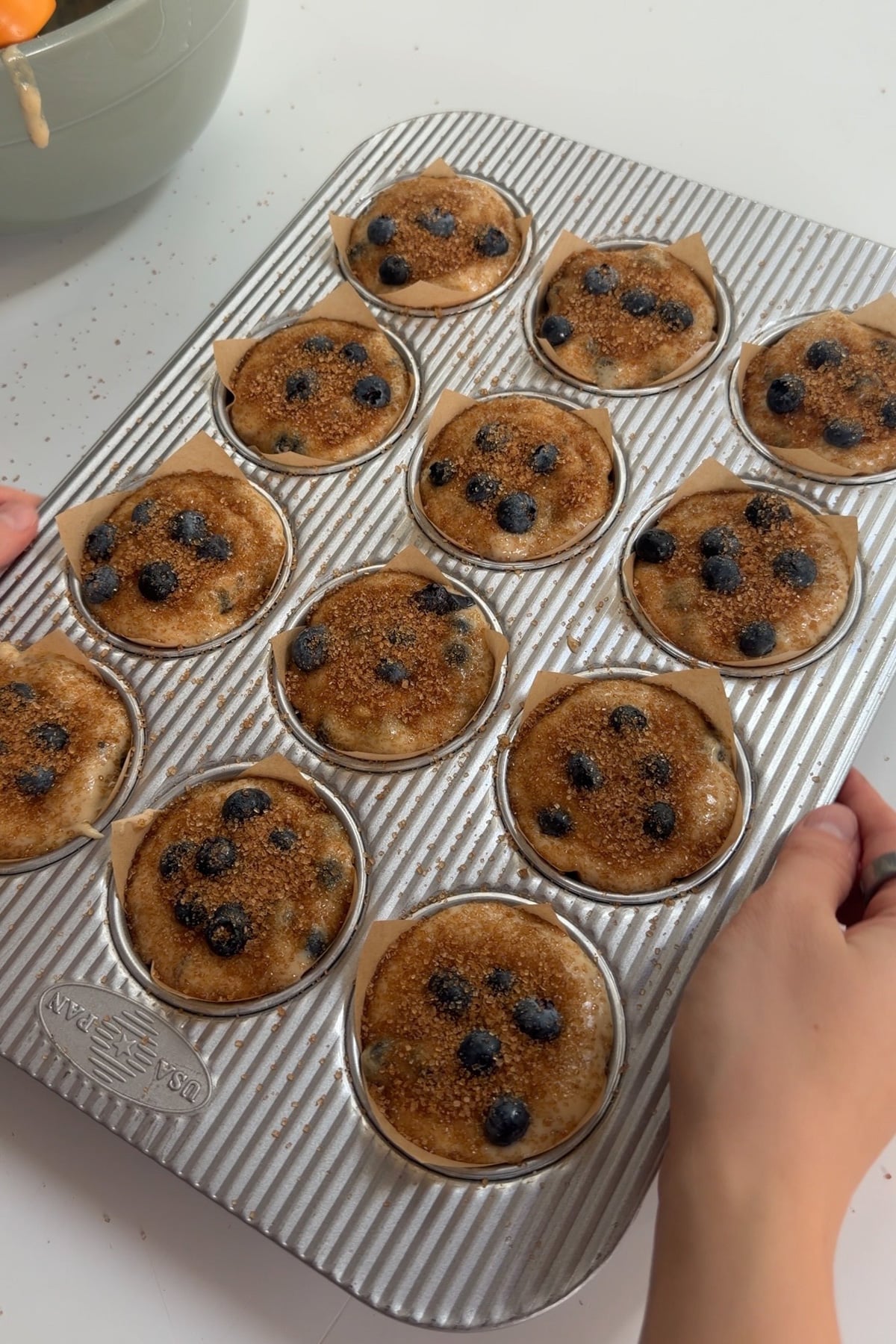 Blueberry banana muffin batter in a 12-muffin pan topped with blueberries and turbinado sugar before baking.